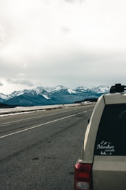 A tranquil road stretches into the distance towards majestic snow-capped mountains under a cloudy sky. A vehicle is partially visible on the right, with a sticker on the window depicting a mountain and the word 'Adventure.' Sparse patches of snow cover the ground beside the road.