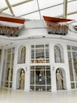 A high-end fashion boutique storefront featuring mannequins adorned in luxurious, detailed garments. The elegant display is set within an architectural facade with large glass panes and decorative arches. Above, flower arrangements with vibrant red and orange flowers add a touch of freshness and color.