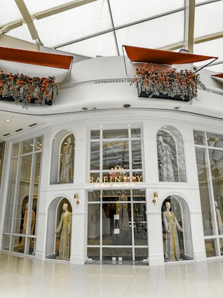 A high-end fashion boutique storefront featuring mannequins adorned in luxurious, detailed garments. The elegant display is set within an architectural facade with large glass panes and decorative arches. Above, flower arrangements with vibrant red and orange flowers add a touch of freshness and color.