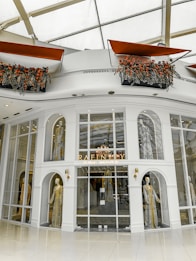 A high-end fashion boutique storefront featuring mannequins adorned in luxurious, detailed garments. The elegant display is set within an architectural facade with large glass panes and decorative arches. Above, flower arrangements with vibrant red and orange flowers add a touch of freshness and color.
