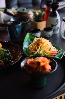 A dining setup featuring various dishes, including a plate of pasta with garnishes, a dish containing what appears to be kimchi, and cooked greens. The table includes multiple small cups and bottles. Various serving ware with artistic designs are also present, and a few potted plants add a touch of nature to the background.