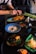 A person is assembling and garnishing various dishes on a table, which include plates with seafood, vibrant salads, and bowls with soup. The table is set with various utensils, ingredients, and a cup, providing a dynamic culinary scene.