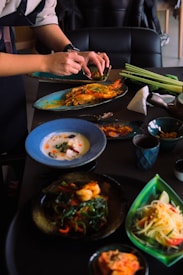 A person is assembling and garnishing various dishes on a table, which include plates with seafood, vibrant salads, and bowls with soup. The table is set with various utensils, ingredients, and a cup, providing a dynamic culinary scene.