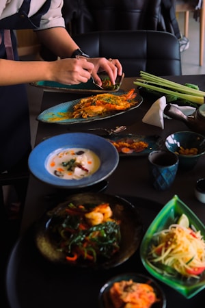 A person is assembling and garnishing various dishes on a table, which include plates with seafood, vibrant salads, and bowls with soup. The table is set with various utensils, ingredients, and a cup, providing a dynamic culinary scene.