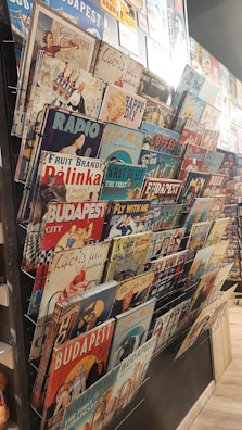 A collection of vintage-style metal signs displayed on a rack. The signs feature various themes, including advertisements, classic imagery, and slogans, with references to Budapest and retro designs.