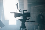 A professional video camera is set up on a tripod in an industrial or studio setting. The camera is positioned facing a bright light source, which creates a dramatic silhouette effect. In the background, there is a person wearing a cap and face mask, standing in a dimly lit area.