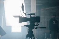 A professional video camera is set up on a tripod in an industrial or studio setting. The camera is positioned facing a bright light source, which creates a dramatic silhouette effect. In the background, there is a person wearing a cap and face mask, standing in a dimly lit area.