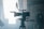 A professional video camera is set up on a tripod in an industrial or studio setting. The camera is positioned facing a bright light source, which creates a dramatic silhouette effect. In the background, there is a person wearing a cap and face mask, standing in a dimly lit area.