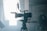 A professional video camera is set up on a tripod in an industrial or studio setting. The camera is positioned facing a bright light source, which creates a dramatic silhouette effect. In the background, there is a person wearing a cap and face mask, standing in a dimly lit area.