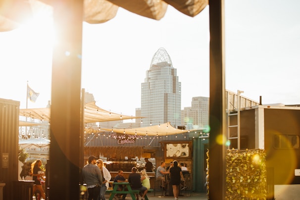 A rooftop bar scene at sunset with several people gathered around tables. The cityscape includes tall buildings and a prominent skyscraper in the background. Warm light creates a cozy and inviting atmosphere with string lights adding to the ambiance.
