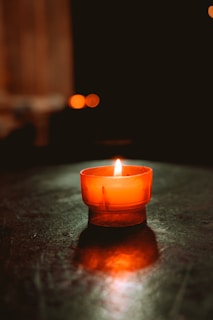 A single, small orange candle is placed on a dark surface, with its flame gently flickering and casting a warm glow. The background is softly blurred, contributing to a serene and intimate atmosphere.