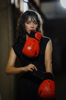 A woman with wet hair stands in a dimly lit space, wearing a black sleeveless top and holding bright red boxing gloves. She appears focused and contemplative, with one glove resting against her chin and the other held by its strap.