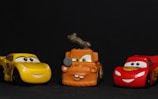 Three toy cars from an animated movie, featuring a yellow race car, an orange tow truck, and a red race car are positioned side by side against a dark background.