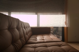 A cozy brown leather sofa with tufted cushions is positioned in a room with soft natural lighting. Behind the sofa, a large window covered by sheer curtains allows a filtered view of the outside, hinting at greenery and relaxed outdoor seating furniture.