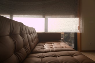 A cozy brown leather sofa with tufted cushions is positioned in a room with soft natural lighting. Behind the sofa, a large window covered by sheer curtains allows a filtered view of the outside, hinting at greenery and relaxed outdoor seating furniture.