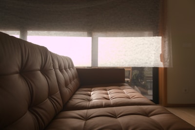 A cozy brown leather sofa with tufted cushions is positioned in a room with soft natural lighting. Behind the sofa, a large window covered by sheer curtains allows a filtered view of the outside, hinting at greenery and relaxed outdoor seating furniture.