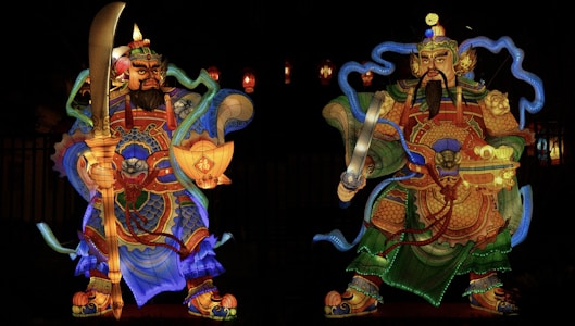 Two illuminated lanterns depicting ancient Chinese warrior figures, adorned in elaborately detailed armor with vivid colors and intricate designs, holding large weapons under a dark night sky.