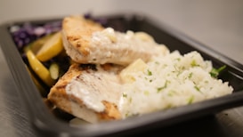 A close-up view of a meal served in a black plastic container. The dish includes grilled chicken topped with a creamy white sauce, a side of white rice garnished with herbs, and a mix of sautéed vegetables including sliced yellow bell peppers and purple cabbage.