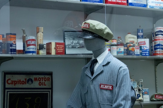 A mannequin dressed as a vintage gas station attendant stands in front of a display filled with old motor oil cans and advertisements. The mannequin wears a gray uniform with an 'Atlantic' patch and a matching cap. The shelves behind contain various automotive products, including Firestone and Capitol brands.