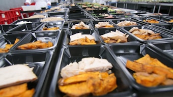 Rows of black meal prep containers are filled with cooked sweet potatoes and slices of turkey, arranged neatly. The containers are placed on a large surface in what appears to be a commercial kitchen setting.