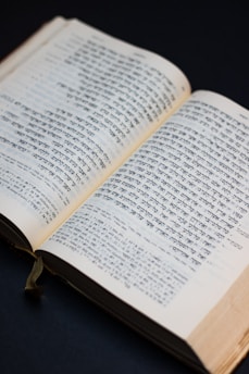 A book is open to display columns of text written in Hebrew script. The pages are slightly yellowed, indicating age or frequent use. A dark background contrasts with the lighter pages, and a green ribbon bookmark is visible along the spine.