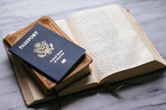 A United States passport is placed on top of a closed leather-bound book, next to an open book with text in a foreign language on a marble surface.