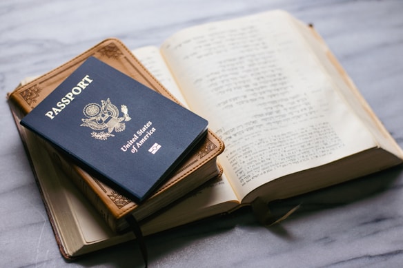 A United States passport is placed on top of a closed leather-bound book, next to an open book with text in a foreign language on a marble surface.