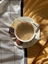 A cup of coffee sits on a saucer adorned with decorative cutouts, accompanied by chocolates and a wrapped golden treat. The setup is placed on a surface covered with two different fabrics, one with a black grid design and the other with yellow polka dots, adding a playful and cozy ambiance.
