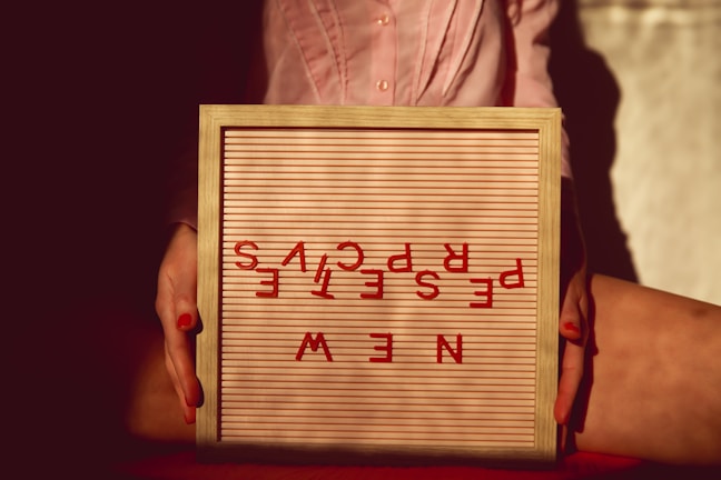 A framed letter board is being held by two hands with red painted nails. The letters on the board are scattered to spell 'NEW PERSPECTIVE' in a playful and artistic, upside-down arrangement. The background is softly lit, adding a warm ambiance to the image.