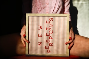 A person holding a framed board with the phrase 'NEW PERSPECTIVES' written in red letters. The board is positioned between their knees, and the person is wearing a pink shirt. The lighting creates a contrast with shadows and highlights.