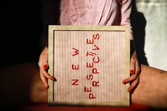 A person holding a framed board with the phrase 'NEW PERSPECTIVES' written in red letters. The board is positioned between their knees, and the person is wearing a pink shirt. The lighting creates a contrast with shadows and highlights.