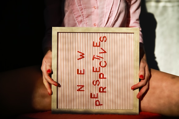 A person holding a framed board with the phrase 'NEW PERSPECTIVES' written in red letters. The board is positioned between their knees, and the person is wearing a pink shirt. The lighting creates a contrast with shadows and highlights.