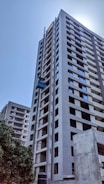 A tall, modern high-rise building under construction with a visible construction elevator. The building has multiple floors with uniform windows and an unfinished appearance. Surrounding the building is a clear blue sky and some greenery at the base.