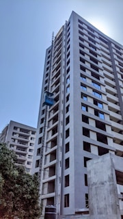 A tall, modern high-rise building under construction with a visible construction elevator. The building has multiple floors with uniform windows and an unfinished appearance. Surrounding the building is a clear blue sky and some greenery at the base.