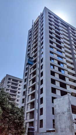 A tall, modern high-rise building under construction with a visible construction elevator. The building has multiple floors with uniform windows and an unfinished appearance. Surrounding the building is a clear blue sky and some greenery at the base.