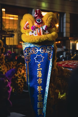 A traditional Chinese lion dance costume in vibrant colors is featured prominently. The lion head is adorned with bright red, pink, and yellow hues, including intricate patterns and decorations. The body banner is a deep blue with gold and yellow Chinese characters, surrounded by intricate design elements. The image is set in an urban environment with blurred backgrounds suggesting a nighttime setting.