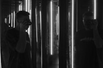 A person is holding a camera in front of mirrors, creating multiple reflections. The lighting is dim with vertical light strips illuminating the scene, adding a dramatic effect.