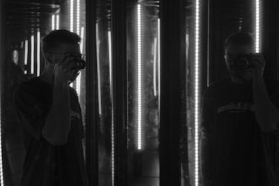 A person is holding a camera in front of mirrors, creating multiple reflections. The lighting is dim with vertical light strips illuminating the scene, adding a dramatic effect.