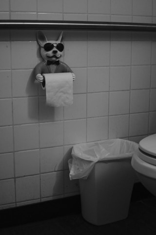 A tiled bathroom wall features a quirky toilet paper holder shaped like a dog wearing sunglasses and a bow tie, holding a roll of toilet paper. Below the holder is a small trash can with a plastic liner, next to part of a visible toilet seat.