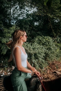 A young woman with long hair, wearing sunglasses and a white top with green pants, is riding a horse through a dense, green forest. She is holding the reins and appears to be calmly enjoying the ride.
