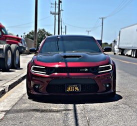 A sleek, modern car with a dark red and black color scheme is parked on the side of an urban street. The license plate reads 'JASS 94'. In the background, large trucks are visible on the road and parked to the side. Utility poles and power lines stretch along the road, under a clear blue sky.