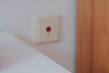 A blurry image showing a white surface in the foreground with a wooden panel on the right. There is a white wall socket or switch with a red button on the wall in the background.