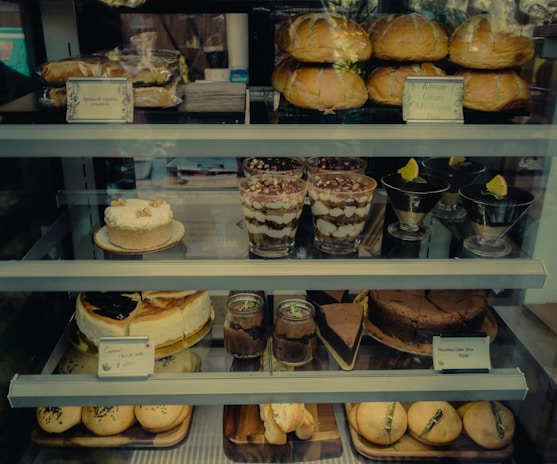 An assortment of baked goods and desserts displayed on shelves, including bread rolls, cakes, tiramisu in glasses, chocolate desserts in dishes, and pastries with labels indicating their names and prices.