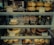 An assortment of baked goods and desserts displayed on shelves, including bread rolls, cakes, tiramisu in glasses, chocolate desserts in dishes, and pastries with labels indicating their names and prices.