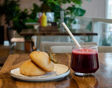On a wooden table, there are two freshly baked buns on a plate, garnished with a chili pepper. Adjacent to the plate is a clear glass filled with a dark red juice, complete with a white straw. In the background, the setting appears to be a cozy cafe with green plants and blurred furniture.