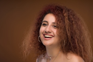 A woman with curly hair and a beaming smile, wearing hoop earrings and a necklace, positioned against a warm brown background.