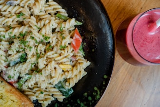 A plate of creamy pasta salad garnished with parsley, featuring colorful vegetables like red peppers and zucchini. Accompanied by a slice of garlic bread, all set on a dark plate. Beside the plate is a glass filled with a vibrant pink smoothie, complete with a straw. Everything is placed on a wooden table, creating a rustic dining ambiance.