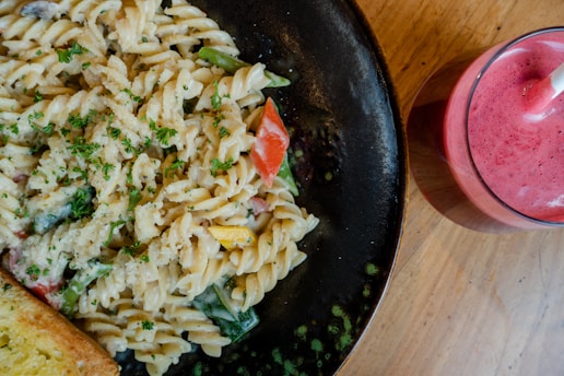 A plate of creamy pasta salad garnished with parsley, featuring colorful vegetables like red peppers and zucchini. Accompanied by a slice of garlic bread, all set on a dark plate. Beside the plate is a glass filled with a vibrant pink smoothie, complete with a straw. Everything is placed on a wooden table, creating a rustic dining ambiance.
