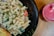 A plate of creamy pasta salad garnished with parsley, featuring colorful vegetables like red peppers and zucchini. Accompanied by a slice of garlic bread, all set on a dark plate. Beside the plate is a glass filled with a vibrant pink smoothie, complete with a straw. Everything is placed on a wooden table, creating a rustic dining ambiance.