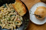 A close-up of a meal on a wooden table. On a black speckled plate, there is creamy pasta with fusilli and mixed vegetables garnished with parsley, accompanied by a slice of toasted garlic bread. To the side, a white plate holds a sesame seed bun with a green chili pepper placed on top of it.